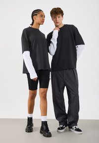 Dwaj modele prezentują nowoczesne, casualowe stroje. Kobieta ma na sobie czarną bluzkę z białymi rękawami oraz szorty kolarskie; mężczyzna nosi czarną bluzkę i luźne szare spodnie.