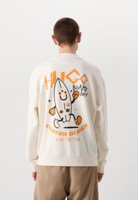 Lys kremfarget sweatshirt med et stort grafisk design av en tegneserie surfebrettkarakter på baksiden, akkompagnert av sterke svarte og orange tekst.