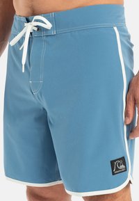 Modré plavky s bílými bočními detaily, vyrobené z lehkého materiálu. Mají stahovací šňůrku a logo nášivku na spodní straně.