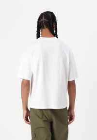 T-shirt en coton blanc avec une coupe décontractée, des manches courtes et un col arrondi, vu de l'arrière. Porté avec un pantalon vert olive.