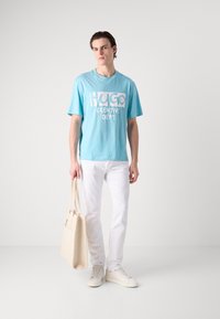Lys blå bomull T-skjorte med hvit grafisk tekst "HUGO CREATIVE DEPT." sammen med hvite jeans og en beige totebag. Enkle hvite sneakers.