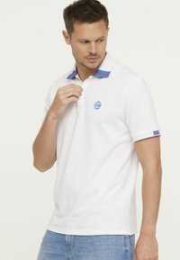 Witte polo shirt met een blauw logo, contrasterende kraag met blauw en rode strepen, korte mouwen met gestreepte accenten, gemaakt van lichtgewicht stof.