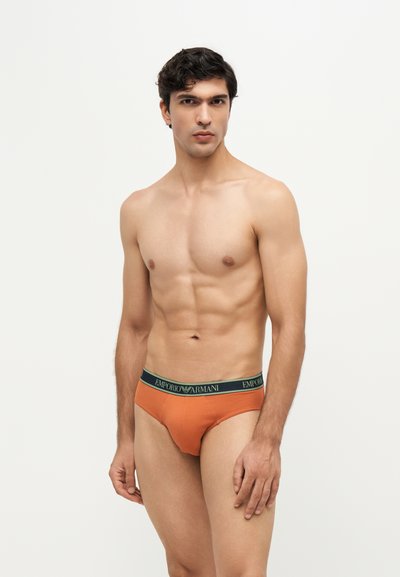 Jeune homme aux cheveux foncés portant un slip orange Emporio Armani, debout devant un fond clair uni.