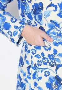 Kukkamallinen kangas sinivalkoisena, jossa on tasku sivussa ja tekstuuri pinta. Hihassa on kynnetty malli.