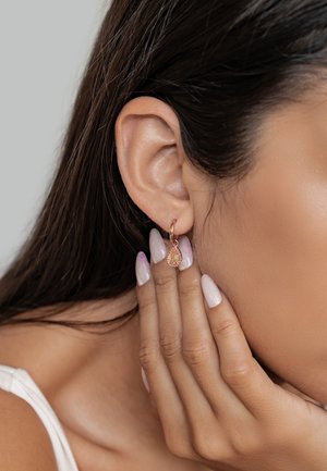 Ohrringe aus Roségold in Form eines Hängers mit einem Tropfenakzent. Die Hand des Models ruht auf dem Gesicht und zeigt hellrosa Nägel mit einem Ombre-Glitzereffekt.