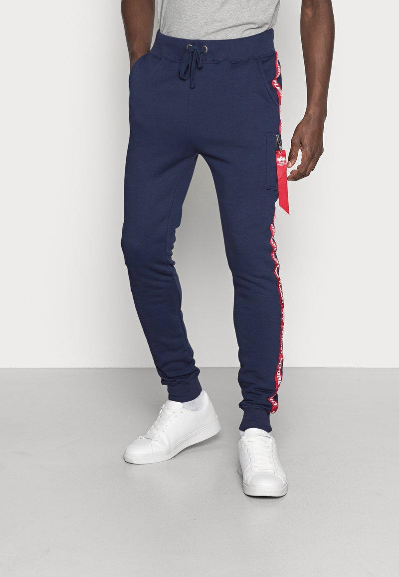 Marineblaue Jogginghosen mit elastischem Bund und Seitentaschen. Mit roten seitlichen Streifen mit Text und einem roten Stoffakzent an der Tasche. Weiße Sneaker.