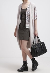 Langer, hellrosa Cardigan mit floralen Details und Spitzenbesatz, getragen über einem dunkelgrünen Kleid. Eine schwarze Handtasche und Ankle Boots runden das Outfit ab.
