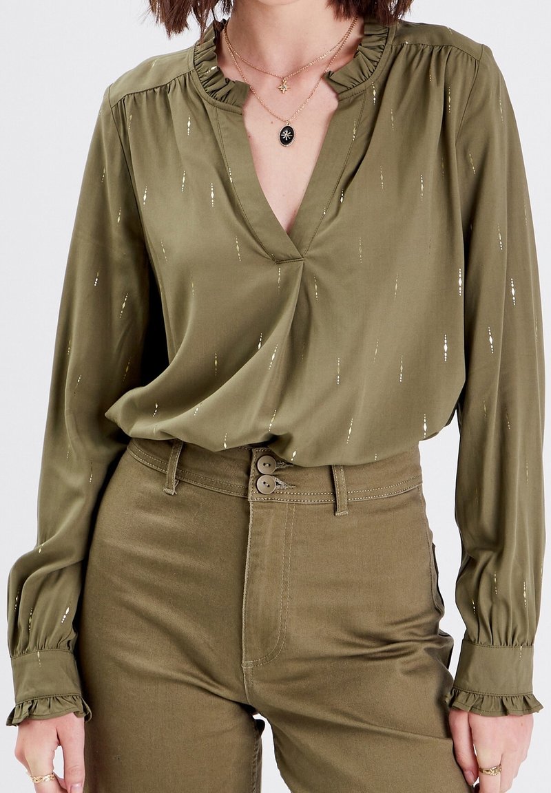 Blouse vert olive avec un décolleté en V et un col à volants, ornée de lignes métalliques discrètes et de manches longues avec des poignets à volants.
