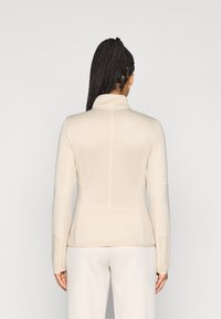 Veste de performance beige à manches longues avec un col montant, des panneaux texturés sur les manches et des détails de design sans coutures à l'arrière.