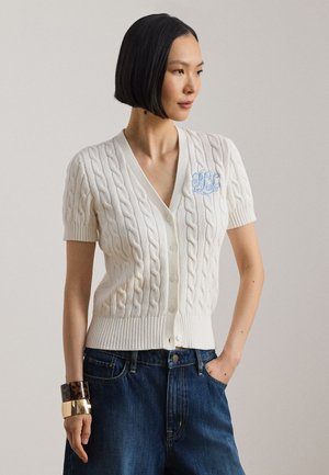 Kobieta o krótkich czarnych włosach, ubrana w biały, krótki sweter na guziki z niebieską haftowaną dekoracją i ciemnoniebieskie dżinsy, stojąca z jedną ręką w kieszeni.