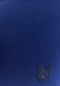 Marineblått stoff med en glatt tekstur, med en liten svart brodert fuglelogo nederst i høyre hjørne.