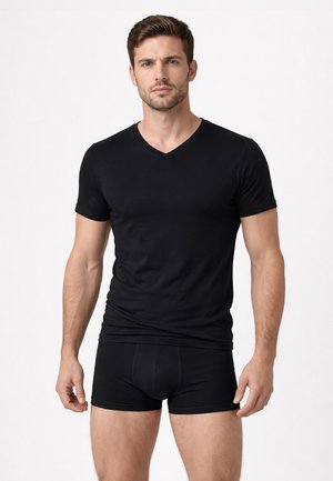 Uomo che indossa una T-shirt nera aderente a scollo a V e boxer neri, in piedi contro uno sfondo chiaro uniforme.