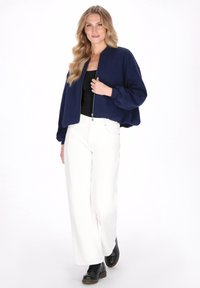 Donkerblauwe bomberjack met ritssluiting, geplooide mouwen en een losse pasvorm, gecombineerd met witte wijd uitlopende broek en zwarte laarzen.