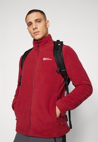 Raudonas flisinis džemperis su aukštu apykakle ir priekiniu užtrauktuku. Papildytas šoninėmis kišenėmis ir Jack Wolfskin logotipu ant krūtinės. Užsimautas virš juodos kuprinės.