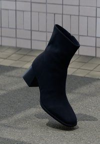 Μαύρο σουέτ μποτάκι μέχρι τον αστράγο με χοντρό τακούνι, ελαφρώς γωνιακό σε επιφάνεια από άσφαλτο κοντά σε τοιχο από πλακάκια.
