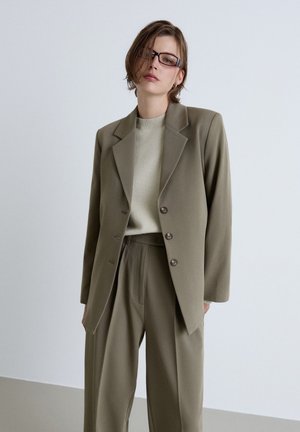 Person mit kurzen Haaren und Brille, die einen olivgrünen Anzug mit einem geknöpften Blazer und passenden weiten Hosen vor einem einfarbigen Hintergrund trägt.