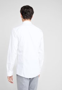 Valkoinen pitkähihaisen paita, joka on valmistettu puuvillasekoitteesta. Siinä on klassinen kaulus, nappi-väli ja kaareva helma. Sujuva pinta.