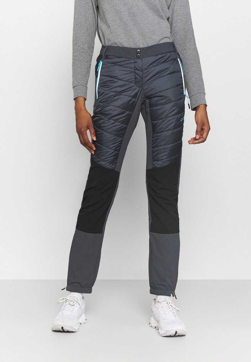 Personne portant un pantalon d'extérieur matelassé noir et gris avec des poches avant zippées bleues et des chaussures de sport blanches, debout sur un fond blanc.