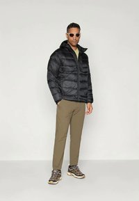 Mand iført solbriller, sort dunjakke, olivengrønne bukser og vandresko, står med hænderne i lommerne mod en ensfarvet baggrund.