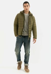 Olivgrüne Pufferjacke mit Kapuze, versehen mit gestepptem Design und schwarzem Reißverschluss. Getragen über einem hellgrauen Pullover und blauen Jeans mit hochgekrempelten Säumen.