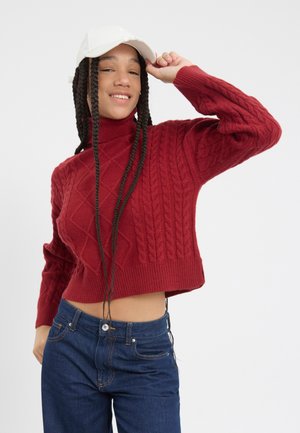 Red cable-knit sweater with a high neck and cropped design, paired with dark blue jeans. The model sports braided hair and a white cap.