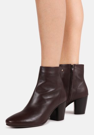Dunkelbraune Leder-Stiefeletten mit abgerundeten Zehen, Blockabsätzen und seitlichen Reißverschlüssen, getragen an nackten Beinen vor weißem Hintergrund.