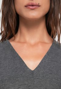 Szürke V-nyakú felső, amely textúrált anyagot mutat, sima nyakkivágással és finom fényléssel, a haj lágyan az vállak körül zuhan.