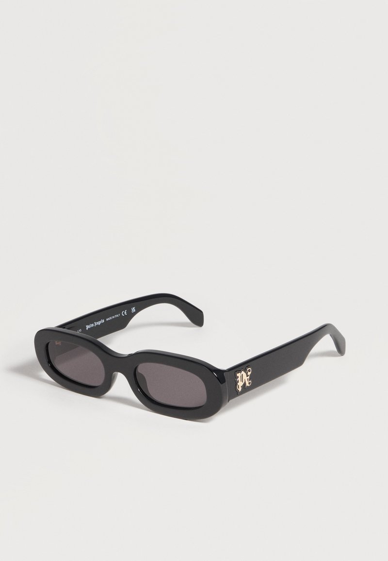 Lunettes de soleil ovales étroites noires avec des verres foncés et un petit logo décoratif doré sur la branche, exposées sur une surface blanche.