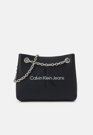 Sac à main Calvin Klein Jeans noir avec une bandoulière en chaîne argentée et un logo "CK" embossé au centre avant.