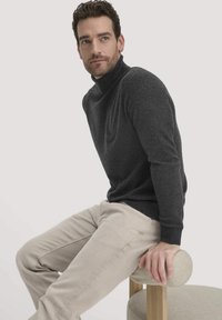 Dunkelgrauer Rollkragenpullover aus weichem Material, kombiniert mit hellbeigen Hosen. Modell sitzt auf einem runden Hocker, die Hände ruhen auf der Sitzfläche.