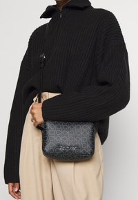 Czarna nylonowa torebka crossbody z wytłoczonym wzorem logo, prostokątny kształt, zamek błyskawiczny i regulowany pasek, trzymana w poziomie w kontrastowym zestawie ze kremowym strojem.