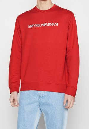 Muž oblečený v červené mikině s dlouhým rukávem s nápisem "EMPORIO ARMANI" v bílé barvě a světle modrých džínách, stojící před jednoduchým pozadím.