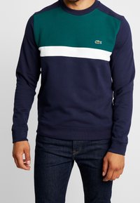 Laivastonsininen collegepaita, jossa vihreät ja valkoiset vaakaraidat, pyöreä pääntie ja pieni logo rinnassa. Materiaali puuvillaa.