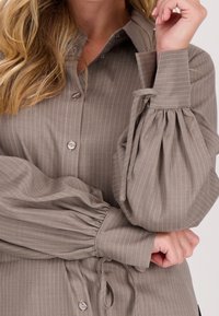 Lichtbruine gestreepte overdubbing met een kraag, voorzien van ballonmouwen met elastische manchetten en strikjes in de taille. Zachte stoftextuur.