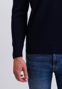 Granatowy sweter dzianinowy z długimi rękawami i ściągaczami, w połączeniu z niebieskimi dżinsami. Zbliżenie ukazuje gładką strukturę i swobodny krój.