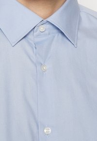 Lichtblauw overhemd met knopen, voorzien van een puntige kraag, met een gladde textuur en zichtbare knopen langs de voorkant.