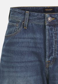 Denim jeans in dark blue with a subtle faded texture. Features a button closure, belt loops, and contrasting yellow stitching.