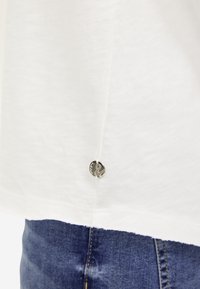 Weißes Baumwoll-T-Shirt mit einem silbernen Logo-Button am Saum, kombiniert mit blauen Jeans, die Textur und Nahtdetails zeigen.