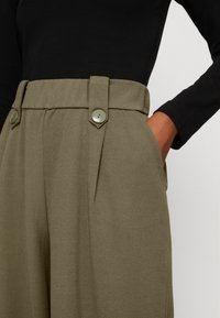 Olivgrüne, hochgeschnittene Hose mit Seitentaschen, vertikalen Falten und zwei großen dekorativen Knöpfen an der Taille. Weiche Textur.