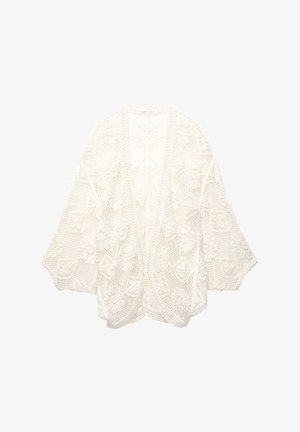 Virkat cardigan av vit bomull med blommönster, öppen front, vida ärmar och strukturerad vävdesign. Har en avslappnad passform.