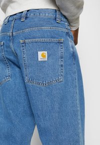 Vaqueros de denim azul claro con un bolsillo trasero cuadrado que lleva una etiqueta blanca y un logo amarillo, mostrando un corte relajado y una textura deslavada.