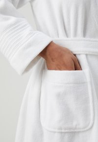 Hvit frottébadekåpe med teksturert erme, knytbelte og lomme foran. Stoffet ser mykt og absorberende ut.