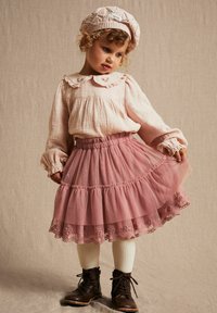 Jupon en tulle rose avec des volants superposés, ourlet en dentelle brodée, associé à un chemisier texturé rose clair et un béret en tricot. Bottes à lacets marron.