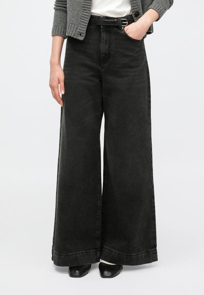 Jean en denim noir à jambes larges avec une taille haute, présentant un tissu uni, des coutures minimales et un ourlet net. Associé à un cardigan gris.