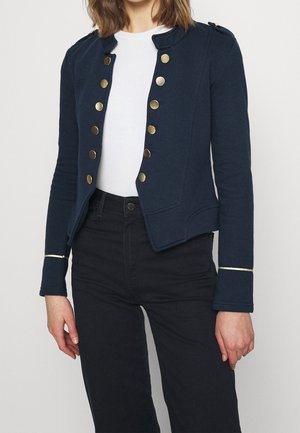 Kobieta ubrana w granatową kurtkę w stylu militarnym z złotymi guzikami, biały top i czarne spodnie z wysokim stanem, stoi z rękami luźno opuszczonymi.