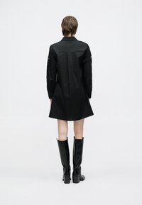 Fekete ingruha hosszú ujjakkal, csipkedíszítéssel; bővülő szegély és strukturált gallér; modell térdig érő fekete csizmával.