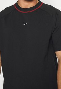 Muž v černém tričku Nike s malým bílým logem swoosh umístěným ve středu a červeným prošitím kolem límce.
