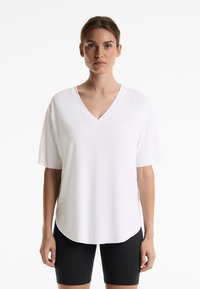 T-shirt blanc à col en V avec des manches courtes, fabriqué en tissu lisse. L'ourlet a une légère courbure, porté avec un short noir ajusté.