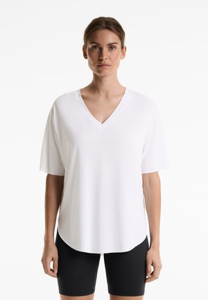 White V-neck T-shirt with short sleeves, made from a smooth fabric. Hem has a slight curvature, worn with black fitted shorts.