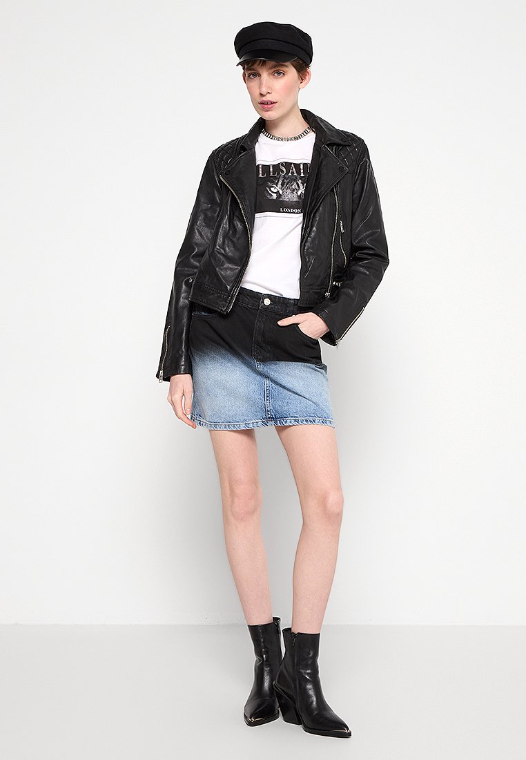 Giacca biker in pelle nera sopra una maglietta grafica bianca, abbinata a una gonna di jeans con effetto ombre e stivali neri alla caviglia.